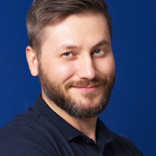 James Newman, a man with short brown hair and a beard, is wearing a dark shirt and stands against a blue background, smiling slightly.