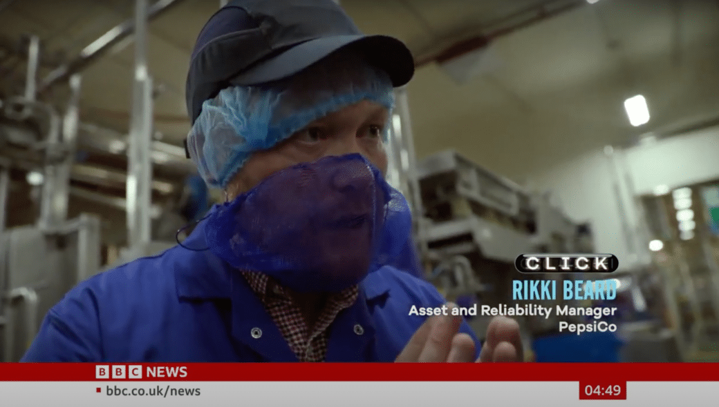 A person identified as an Asset and Reliability Manager for PepsiCo, named Rikki Beard, is speaking inside a factory, wearing a blue hairnet and beard cover. Amid the snack-making sounds, the BBC News ticker is visible at the bottom.