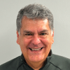 A middle-aged man with gray hair, known as Ed Ballina, smiles against a plain background. He is wearing a dark green zip-up jacket.