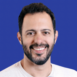A man with a beard and short hair, resembling Gal Shaul, smiles in front of a solid blue background. He is wearing a light-colored shirt.