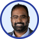 A man with glasses and a beard smiles, wearing a grey blazer and white shirt against a light blue background, epitomizing the spirit of advancing technology.