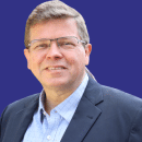A man with glasses, wearing a suit jacket over a blue shirt, reminiscent of Niels Andersen’s classic style, stands against a matching blue background.